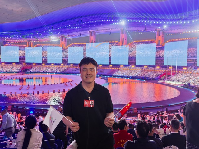 Dr Howard Long inside a stadium at the 15th National Games opening ceremony, holding a small national flag.